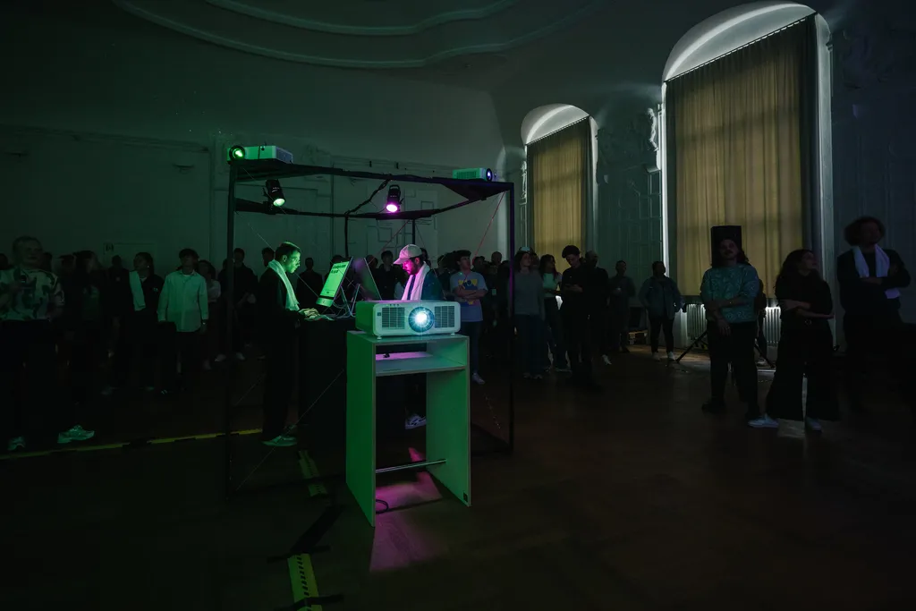 The image depicts an indoor event with a crowd of people watching a person using a computer on a stage illuminated by purple and green lights. The audience appears engaged with the presentation, and the room has a historical decor with large draped windows.