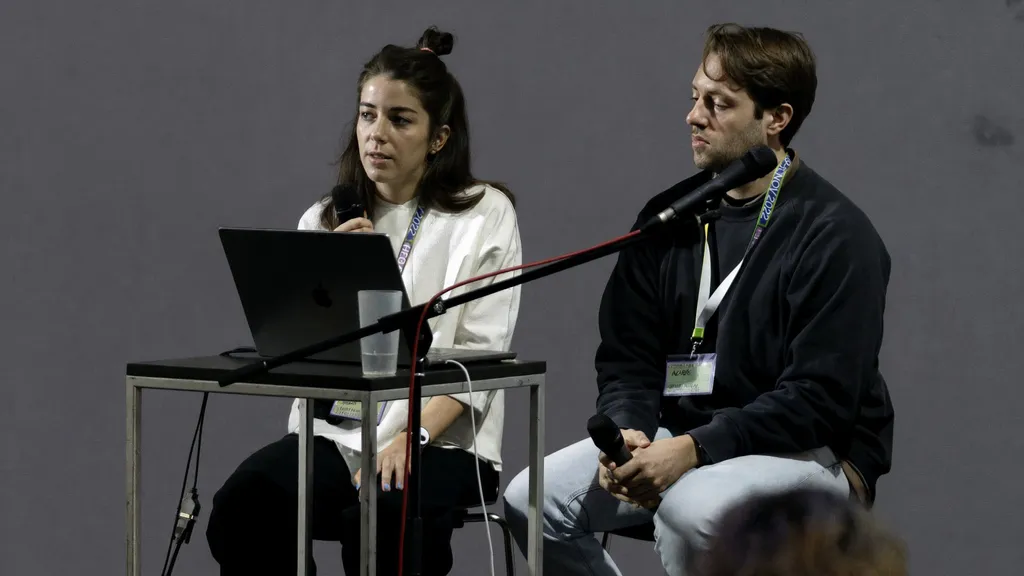 Two people presenting at a conference, one woman with a laptop and a microphone and one man with a microphone, both sitting at a table with lanyards.