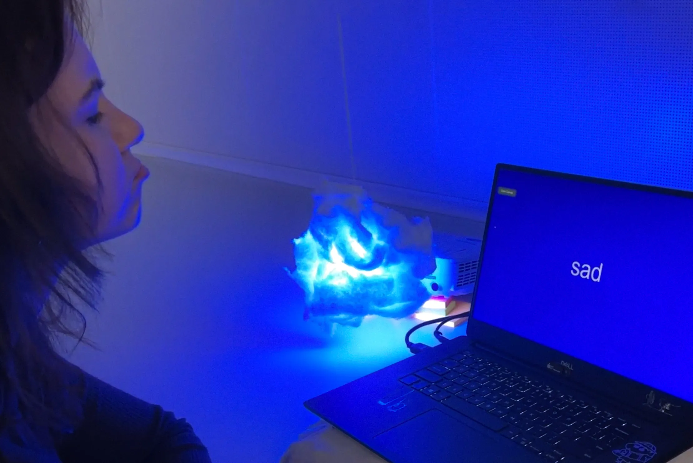 A person looks at a cloud-shaped lamp glowing in deep blue light, placed next to a laptop. The laptop screen displays the word "sad" in white text on a blue background, indicating the detected facial expression. The lamp's colour and intensity respond to the emotion recognised by the system. 
