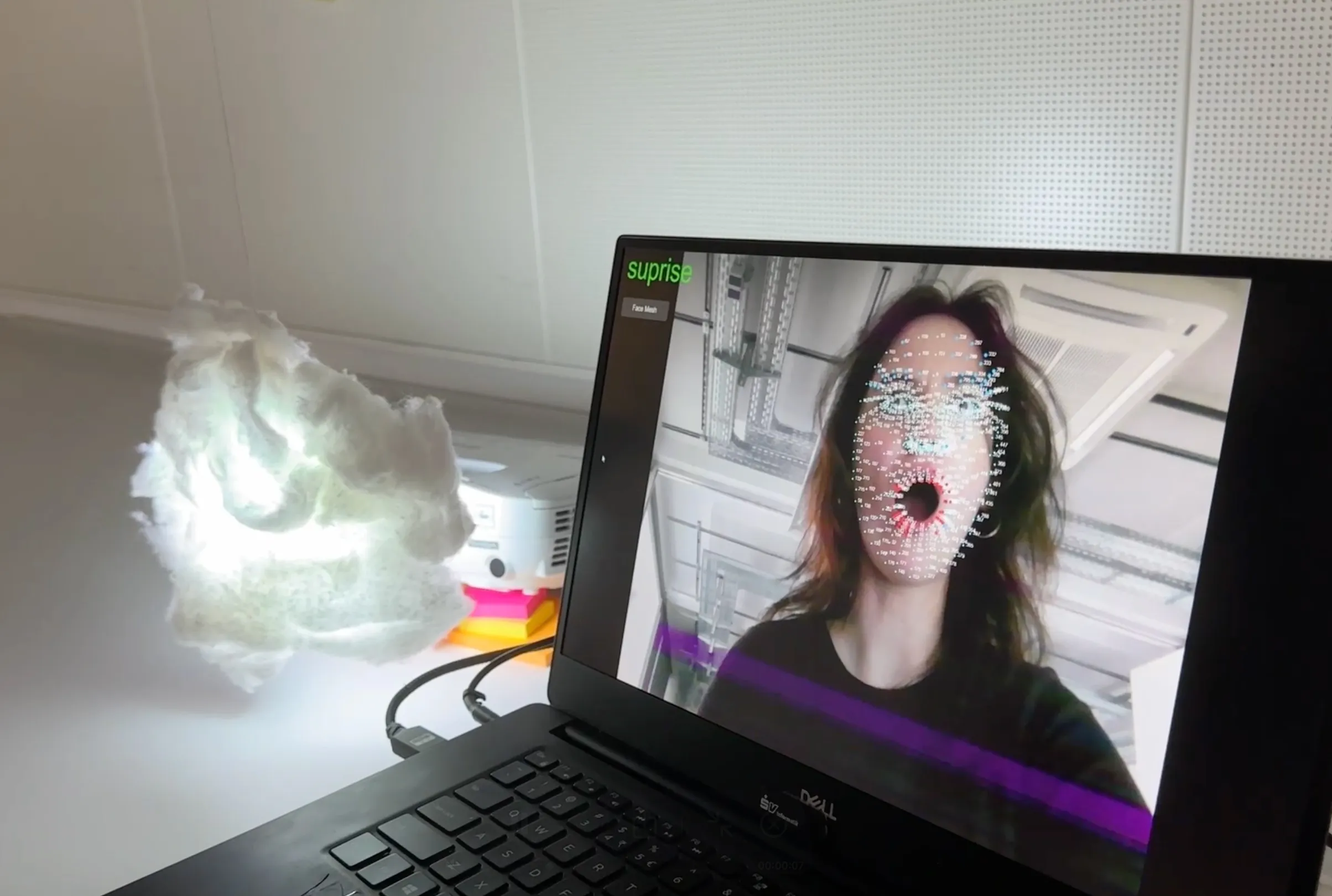 A laptop screen shows a live camera feed of a person's face overlaid with a face mesh — a grid of tracking points used for facial expression recognition. The label "surprise" is displayed in green text in the top left corner. In the background, the cloud-shaped Gloomy lamp emits a soft white glow, ready to respond to the detected emotion